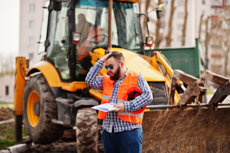 Brutal beard worker man suit construction worker in safety orange helmet, sunglasses against traktor with plan paper at hands.